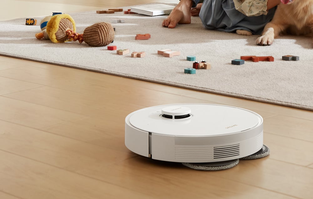biały robot sprzątający mopuje drewnianą podłogę, w tle dywan z porozrzucanymi zabawkami